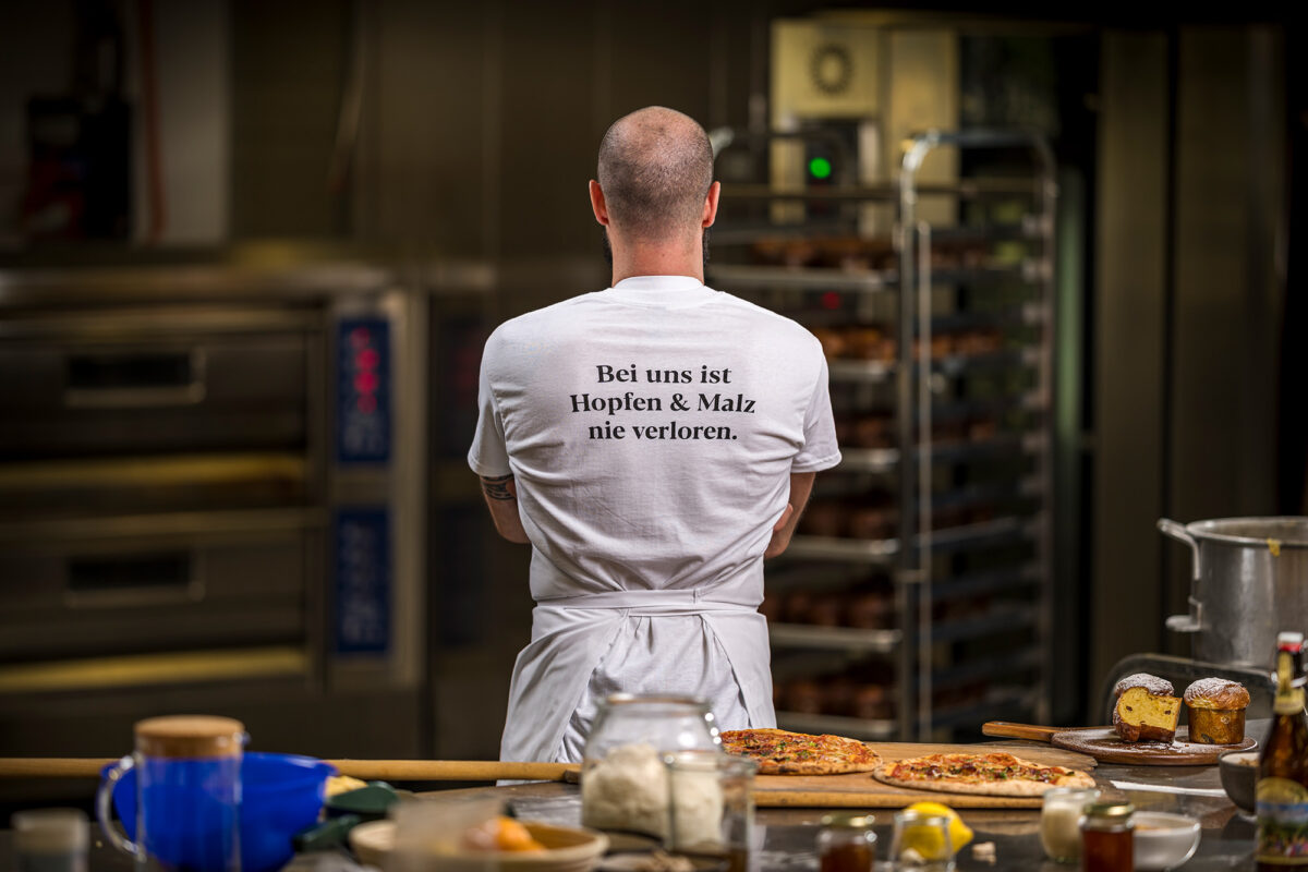 Ein Bäcker in einer Bäckerei trägt ein T-Shirt mit der Aufschrift "Bei uns ist Hopfen & Malz nie verloren", Pizzen und Gebäcke im Vordergrund.