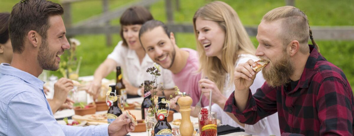 Gruppe junger Leute beim gemeinsamen Essen im Freien, lachend und entspannt am gedeckten Tisch mit Getränken und Snacks in grüner Umgebung