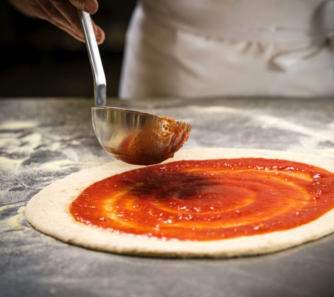 Hand streicht mit einer Kelle frische Tomatensauce auf rohen Pizzateig – Schritt der handwerklichen Pizzazubereitung in der Brauerei.