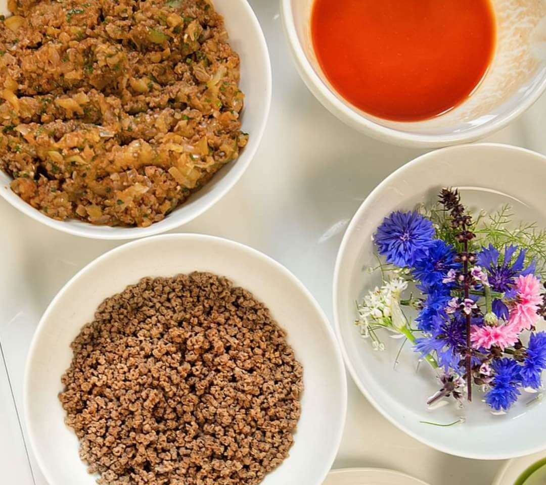 Pflanzliche Zutaten in Schalen: gewürztes Vegi-Hack, getrocknetes Granulat, Tomatensauce und essbare Blüten zur Zubereitung.