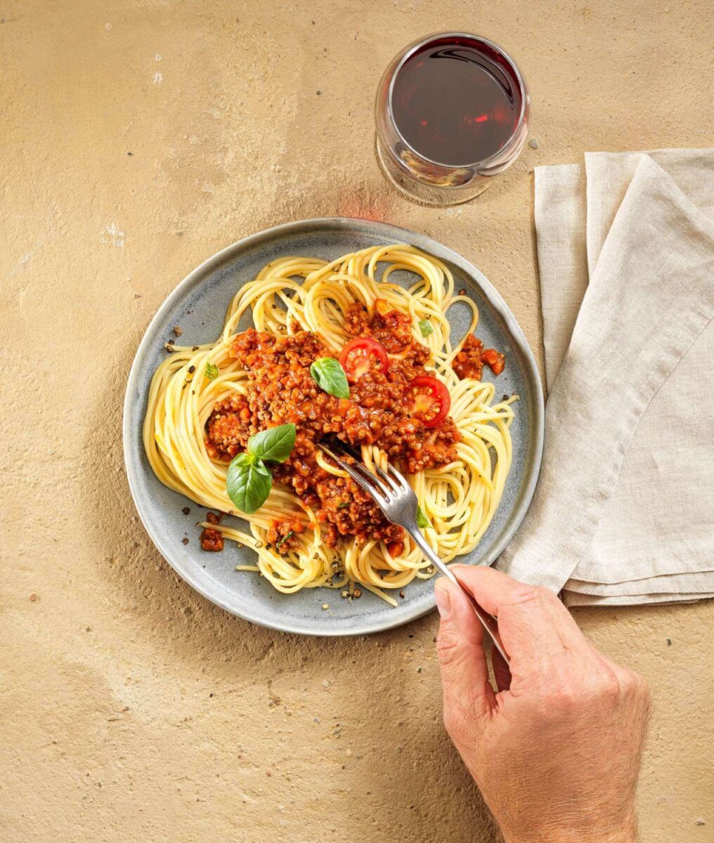 Plant-based Spaghetti Bolognese auf einem Teller mit Tomaten und Basilikum, daneben ein Glas Rotwein und eine Hand mit Gabel.