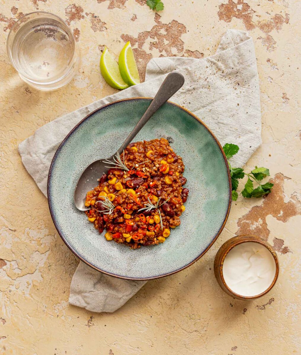 Teller mit Chili sin Carne, serviert mit Mais, Bohnen und Kräutern, dazu Limetten, Wasser und Dip auf hellem Untergrund