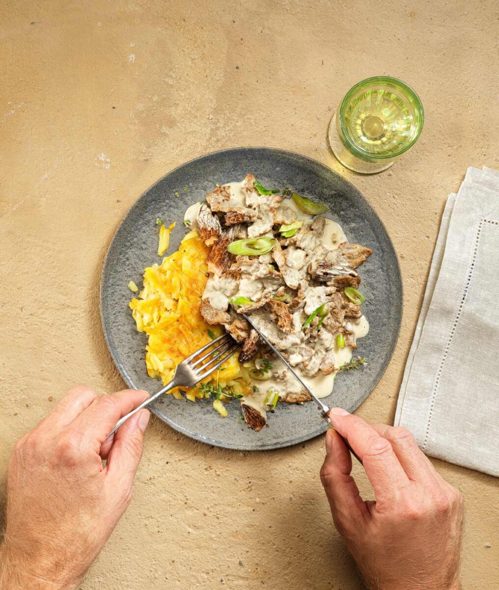 Teller mit pflanzlichem Rahmgeschnetzeltem und Rösti, dazu ein Glas Getränk; Hände schneiden das Gericht auf hellem Untergrund