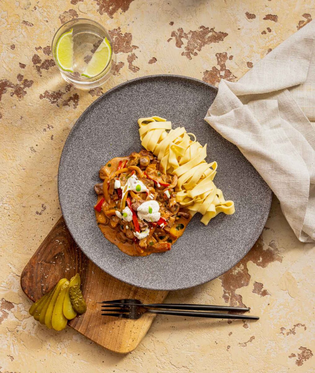 Teller mit pflanzlichem Stroganoff und Bandnudeln auf Holzbrett, dazu Gurken, Wasser mit Limette und helles Tuch