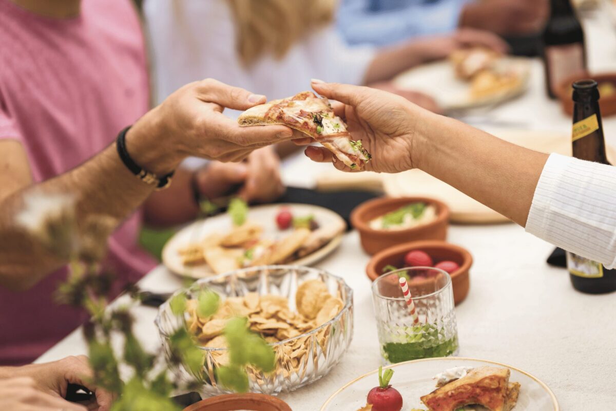 Mehrere Personen sitzen gemeinsam an einem Tisch im Freien und teilen ein Stück Pizza; auf dem Tisch stehen Chips, kleine Schalen mit Gemüse und Dips, Getränke und weiteres Essen.