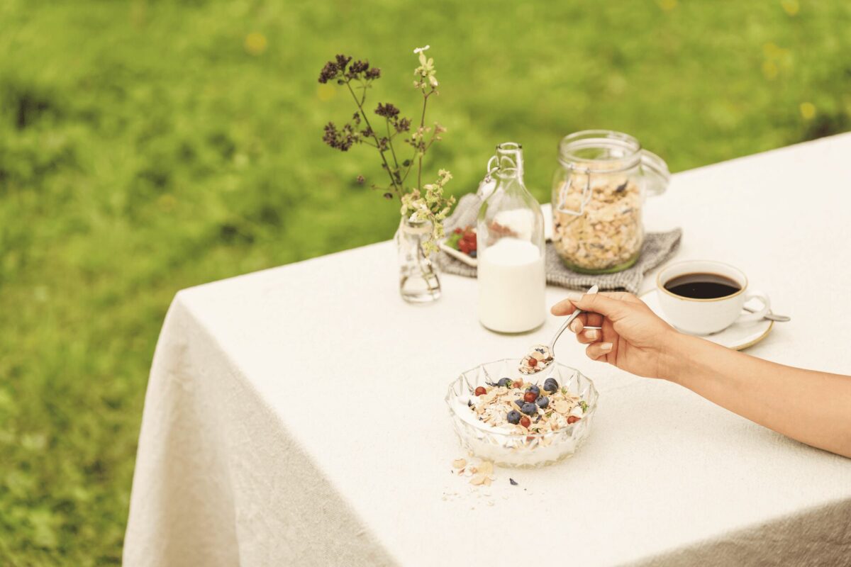 Eine Person isst an einem Tisch im Freien eine Schüssel Müsli mit Beeren; daneben stehen Milch, Granola, Kaffee und ein kleines Blumenarrangement.