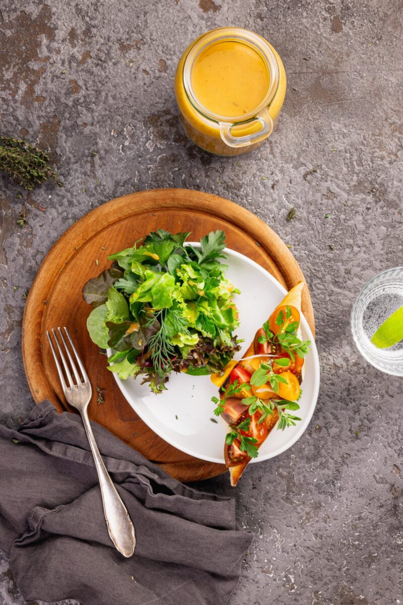 Ein Teller mit frischem gemischtem Salat und einer Tomaten-Bruschetta, serviert auf einem runden Holzbrett; daneben ein Glas Wasser mit Limette, ein Glas mit gelbem Dressing und ein Besteck auf einer dunklen Serviette.